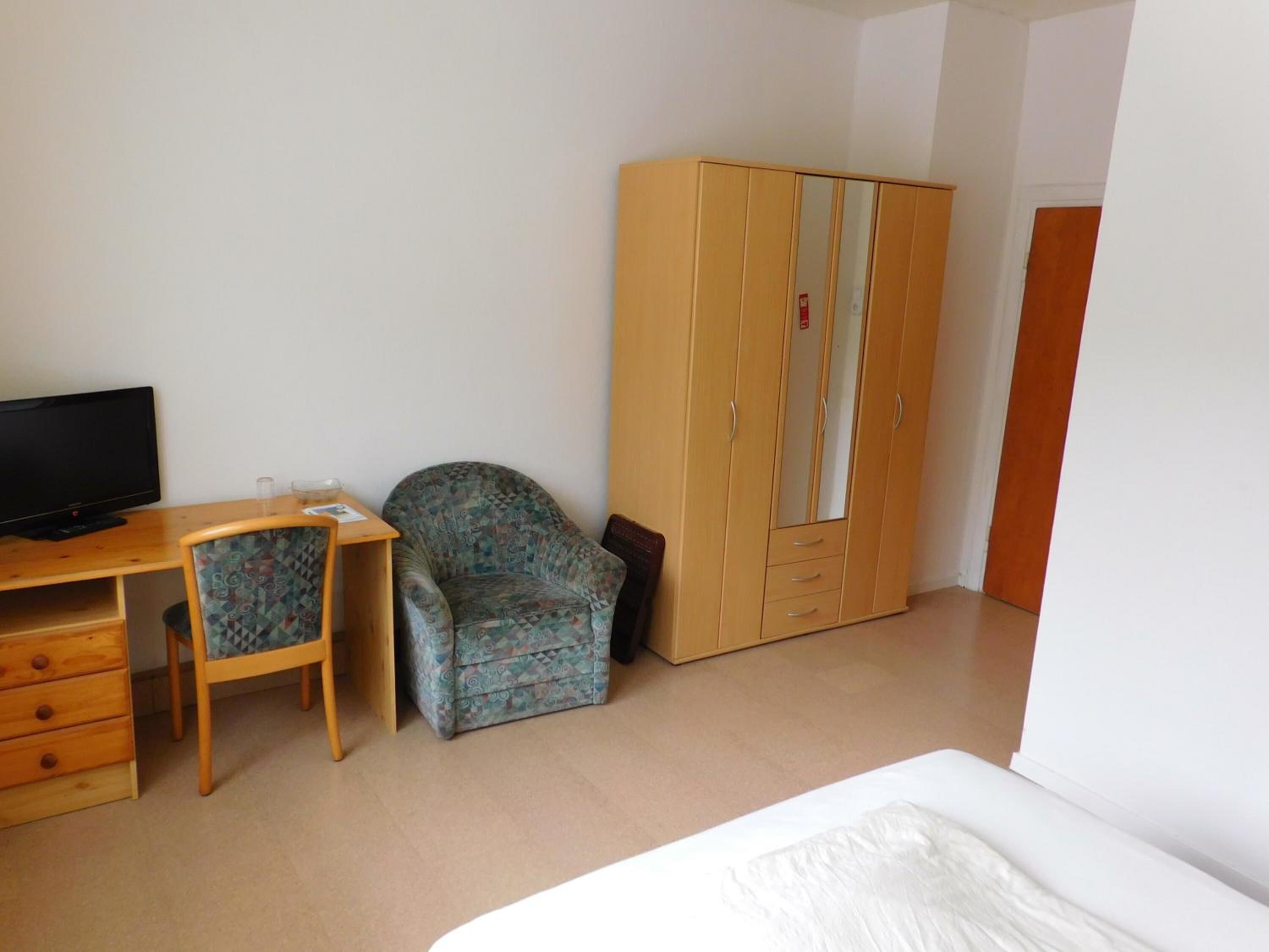 A room with a bed, a desk with a television, a patterned armchair, and a wardrobe.