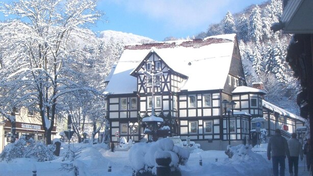 A traditional half-timbered building with a snow-covered roof, surrounded by snow-laden trees and a mountainous backdrop.
