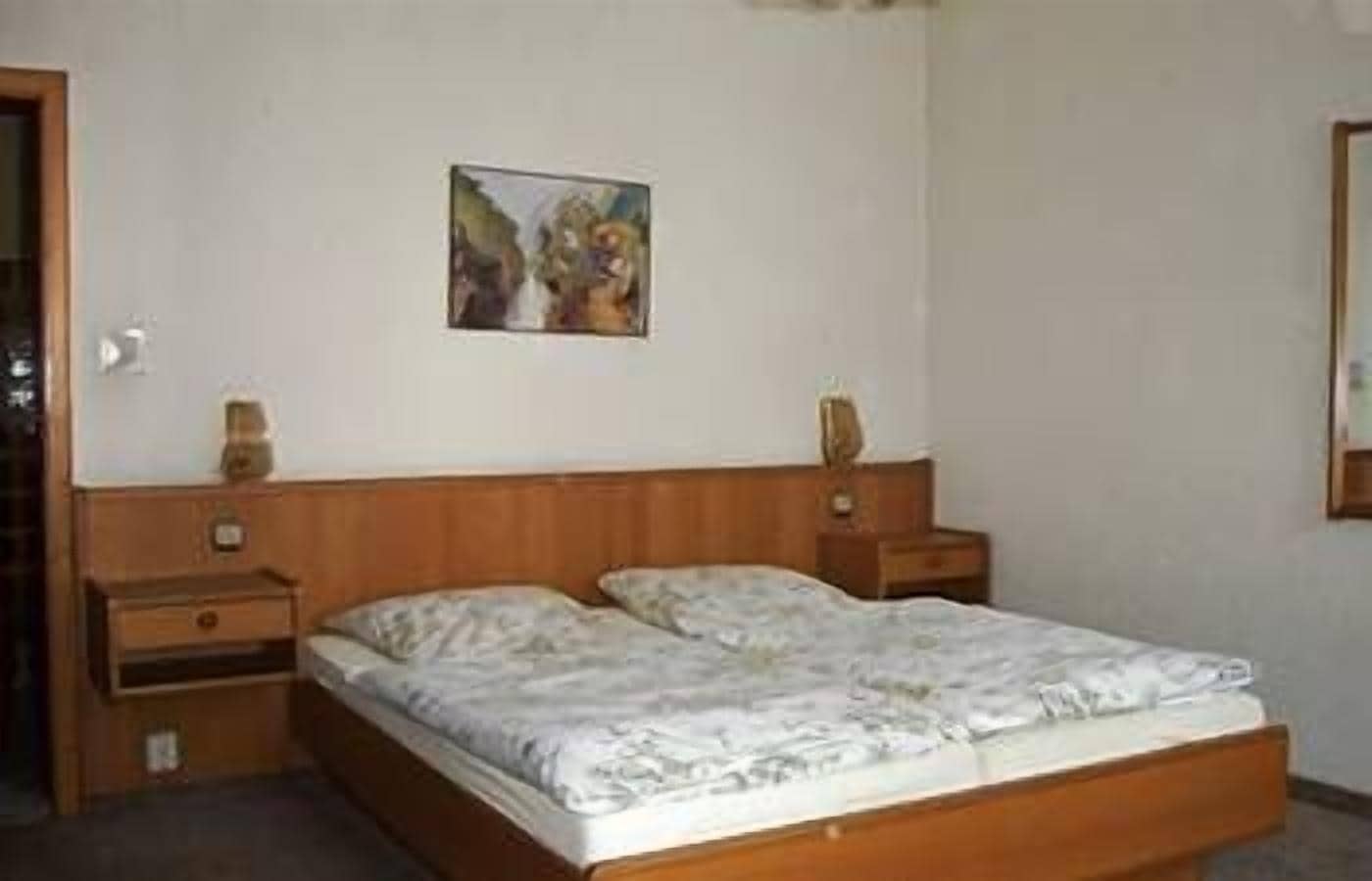 Quarto com estrutura de cama de madeira, mesa de cabeceira com abajur e um quadro na parede.