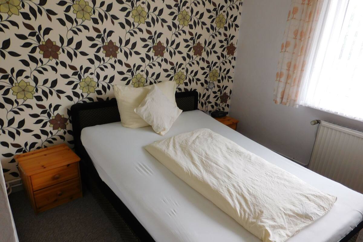 Ein Schlafzimmer mit einem Bett, zwei Nachttischen, einer Holzkassette und einem blumigen Wandmuster.