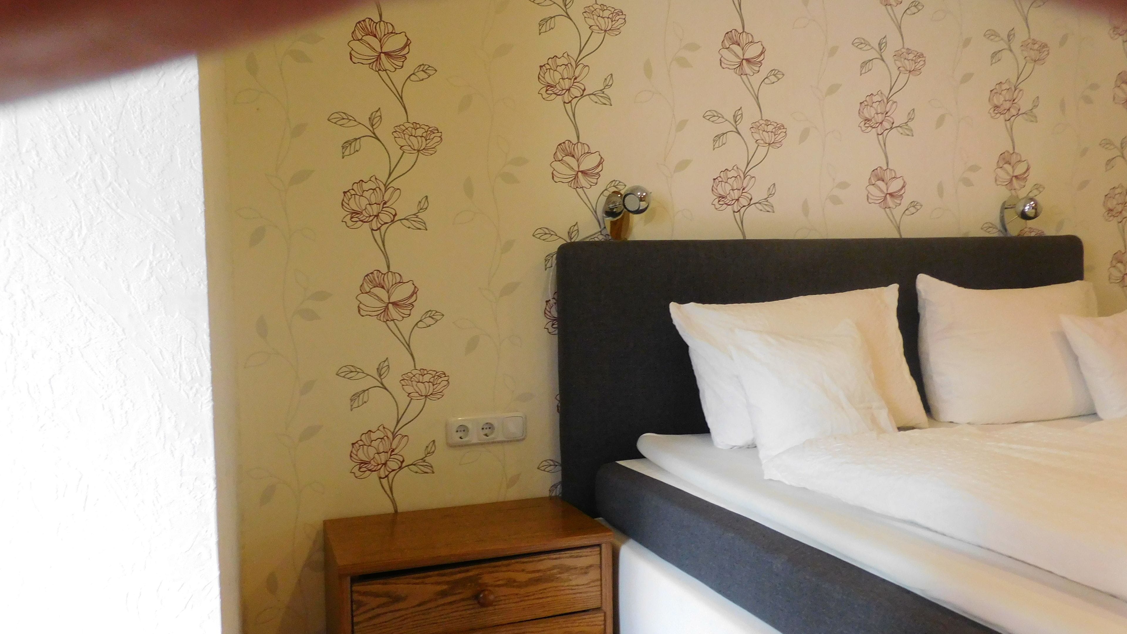 A bedroom with a bed, white pillows, a wooden nightstand, and floral wallpaper.