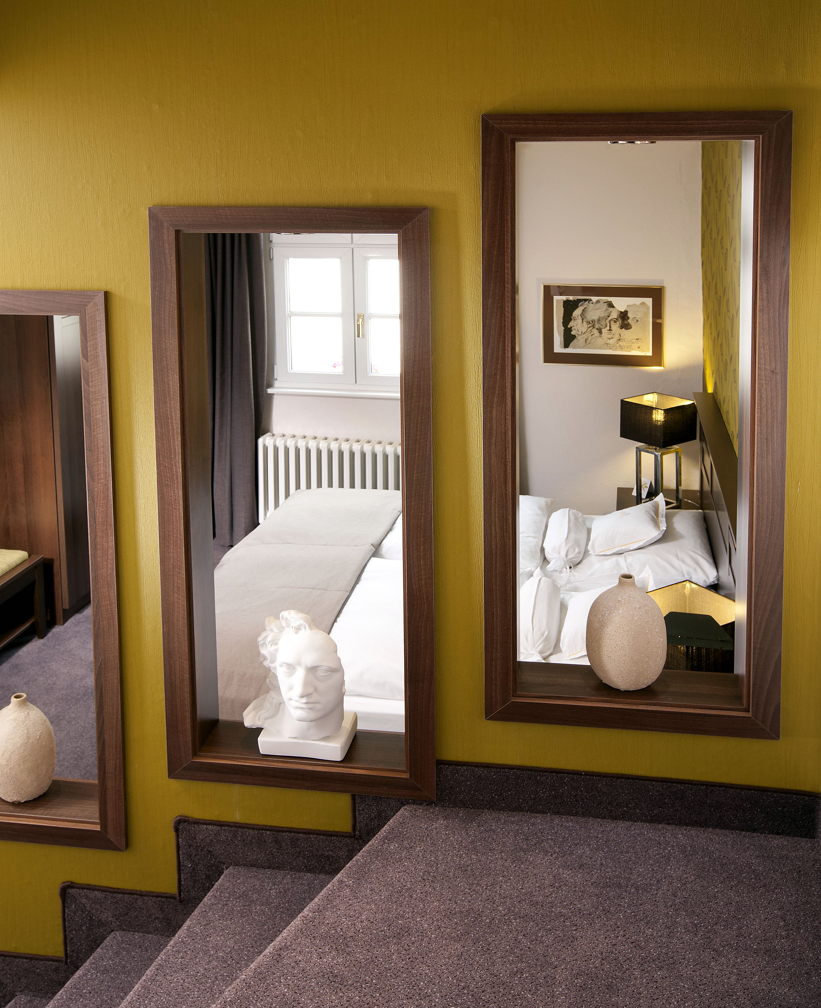 A hotel room with a bed, a bedside lamp, a framed mirror, and a sculpture on the nightstand.