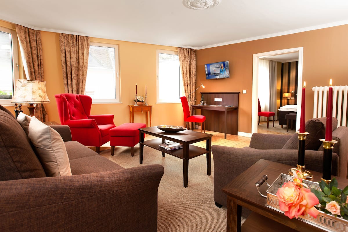 Een woonkamer met een bruine bank, een rode fauteuil en een houten salontafel. Er hangt een televisie aan de muur en op een klein tafeltje staan kaarsen en bloemen.