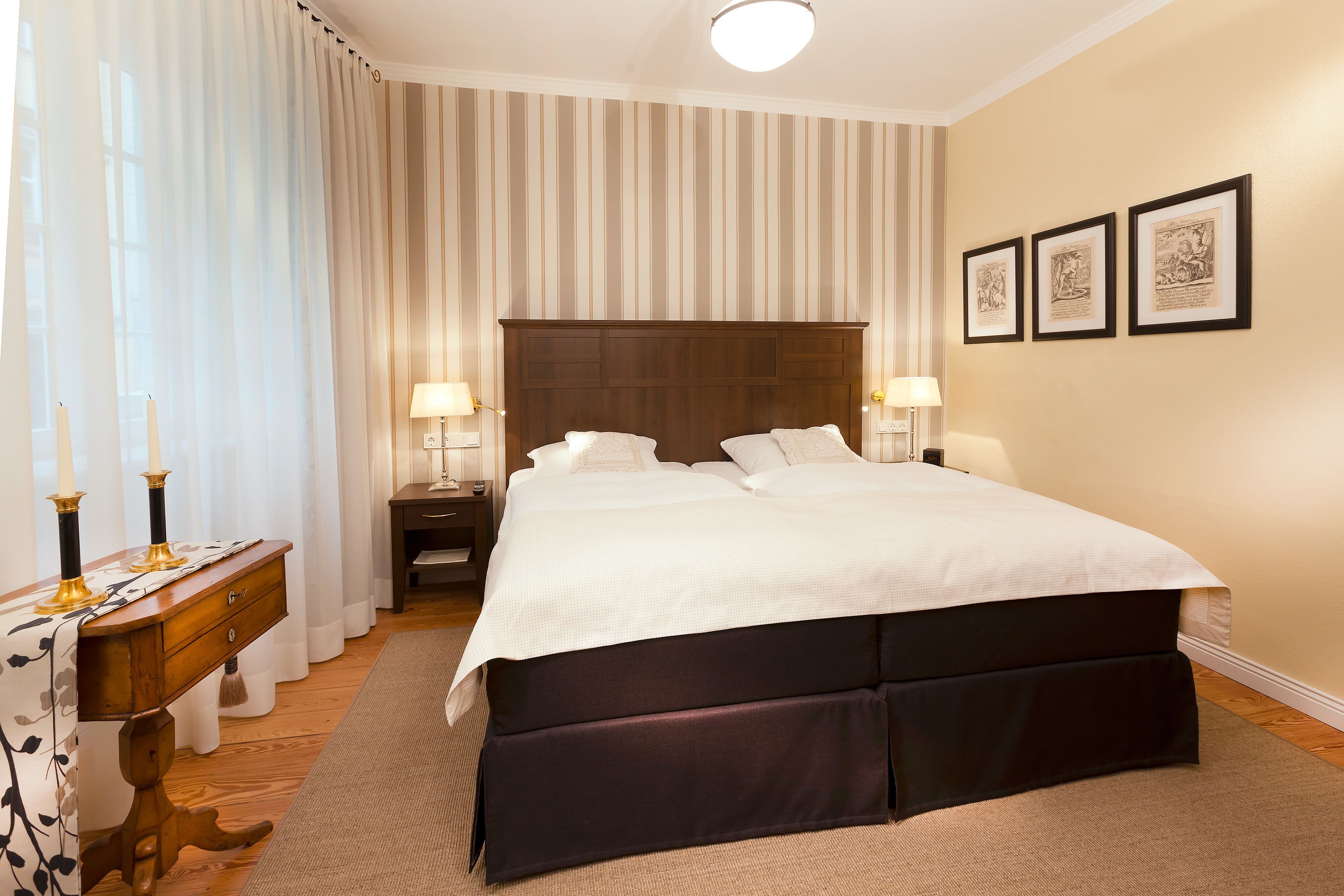 A neatly arranged bedroom with a large bed, two bedside tables with lamps, a wooden dresser, and a large window with curtains.