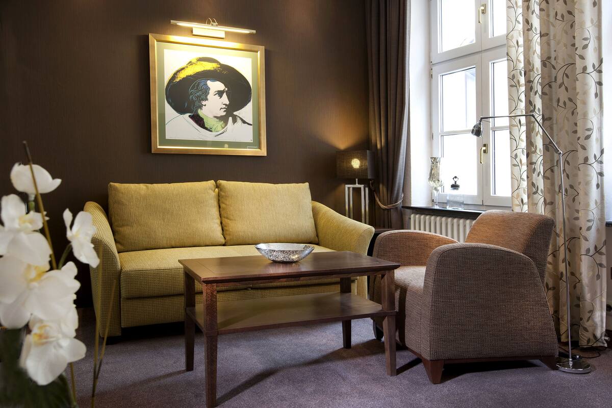 Een kamer met een bank, een stoel, een tafel, een lijst met een portret en een raam met gordijnen.