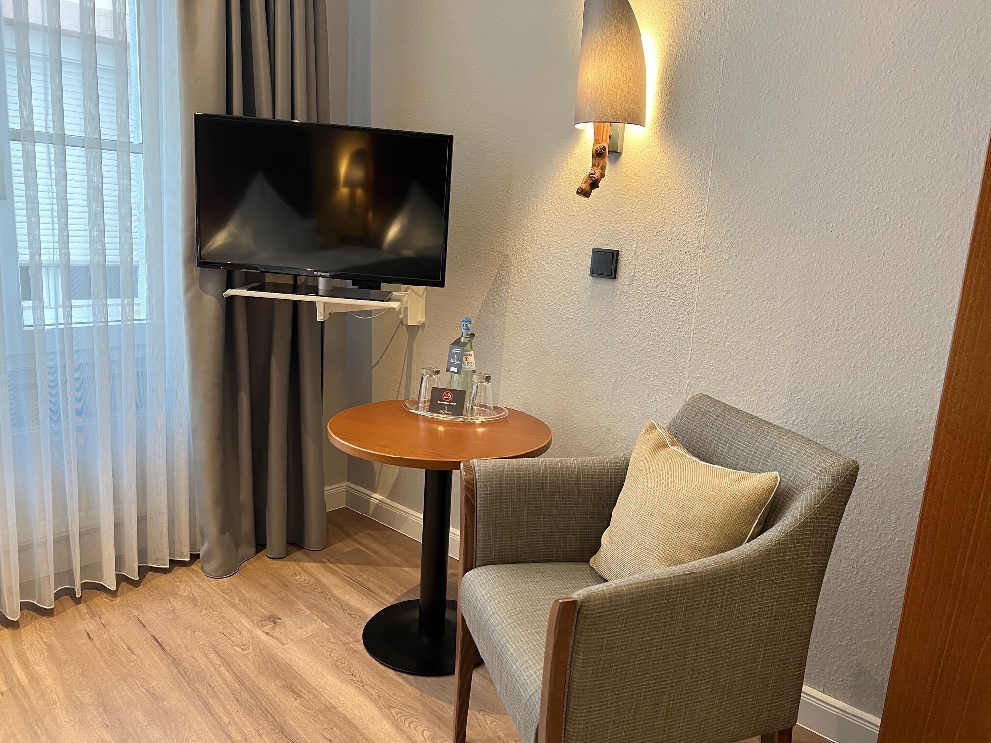 A hotel room with a television on a stand, a small round table with a bottle and glasses, a grey armchair, and a window with curtains.