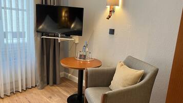 A hotel room with a television on a stand, a small round table with a bottle and glasses, a grey armchair, and a window with curtains.