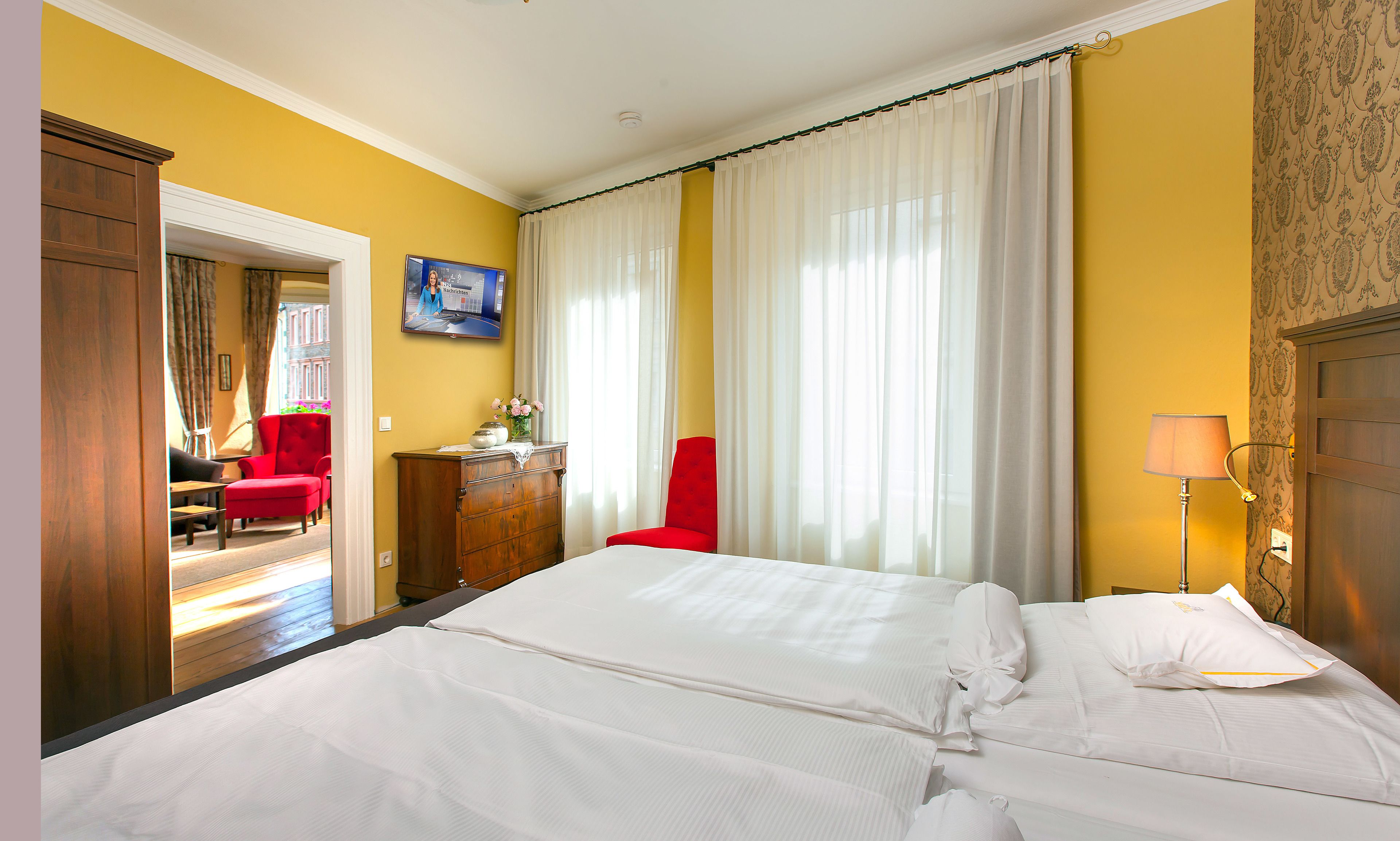 Ein Hotelzimmer mit einem Bett, einem roten Sessel, einer Holzkommode und einem Fernseher an der Wand.