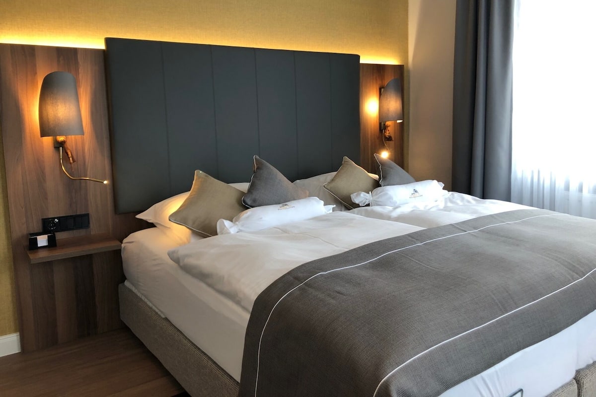 Een hotelkamer met een groot bed, meerdere kussens, een hoofdbord met ingebouwde verlichting, een nachtkastje met een lamp en een raam met gordijnen.