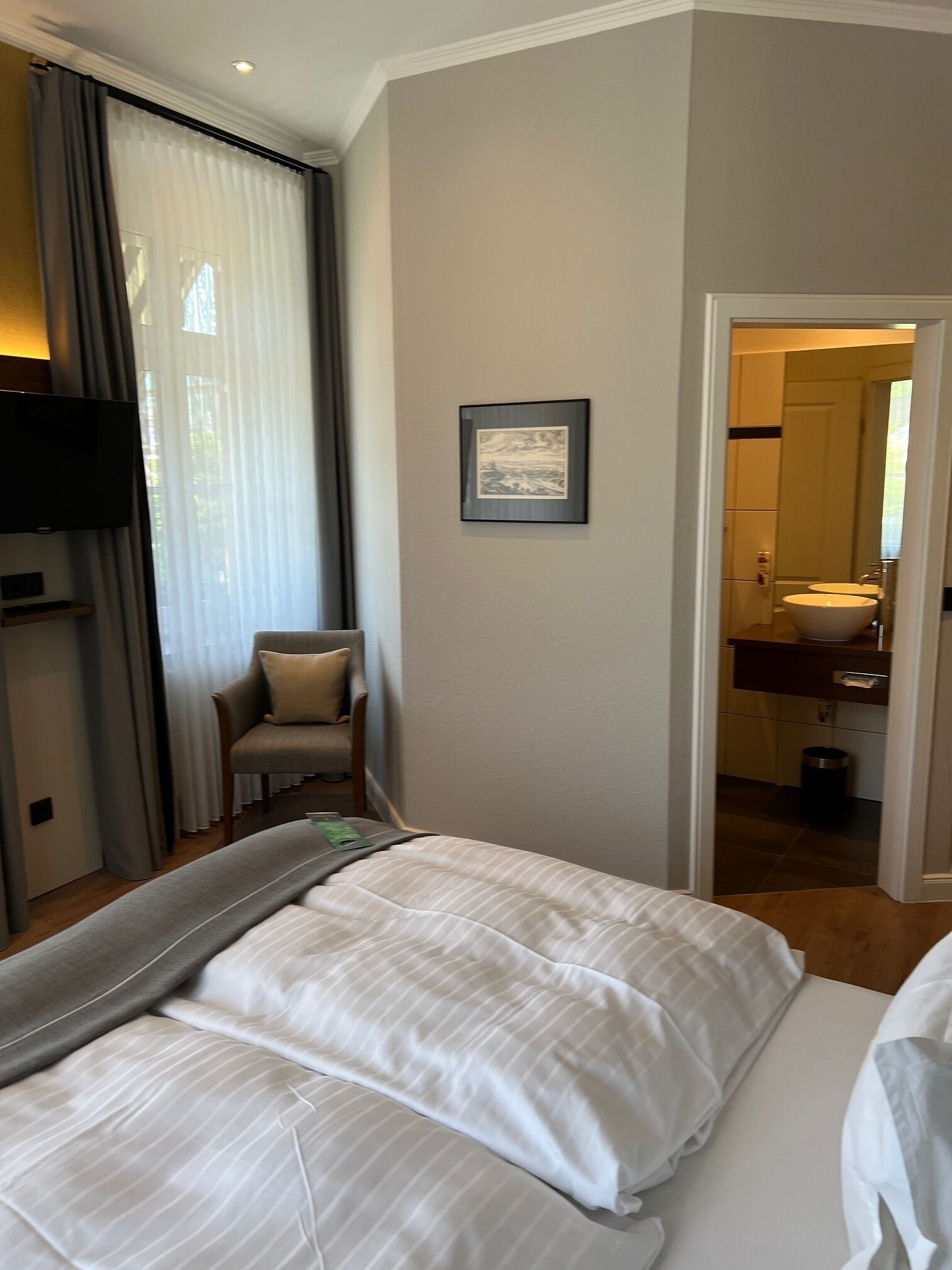 A hotel room with a bed, a chair, a television, a framed picture on the wall, and a bathroom visible through an open door.