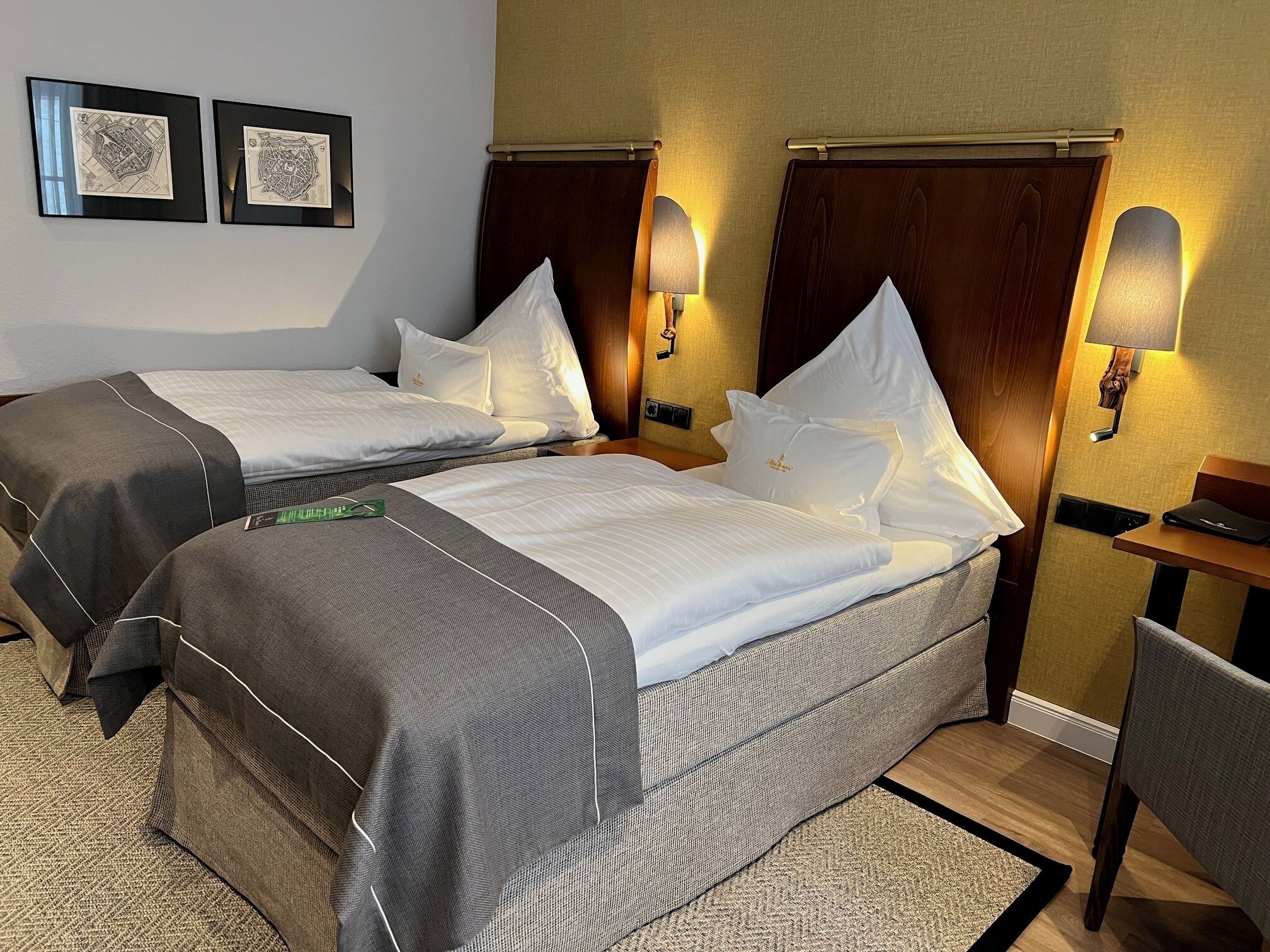 A hotel room with two beds, a desk, and a chair. There are two wall-mounted lamps and two framed maps on the wall.