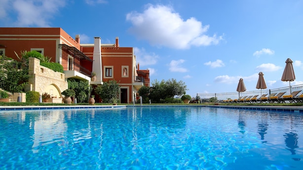 A swimming pool with a clear blue water surface, surrounded by a building with a red facade and a stone wall. - Katalagari Country Suites (Archanes-Asterousia)