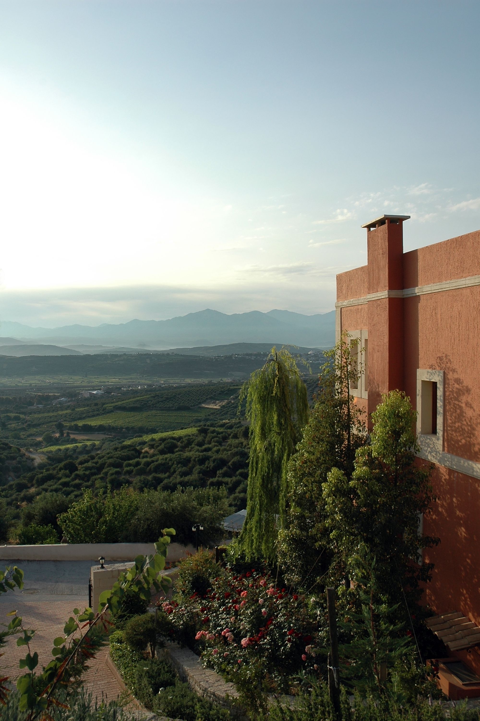 A building with a red facade, a garden with various plants and flowers, and a view of distant mountains.