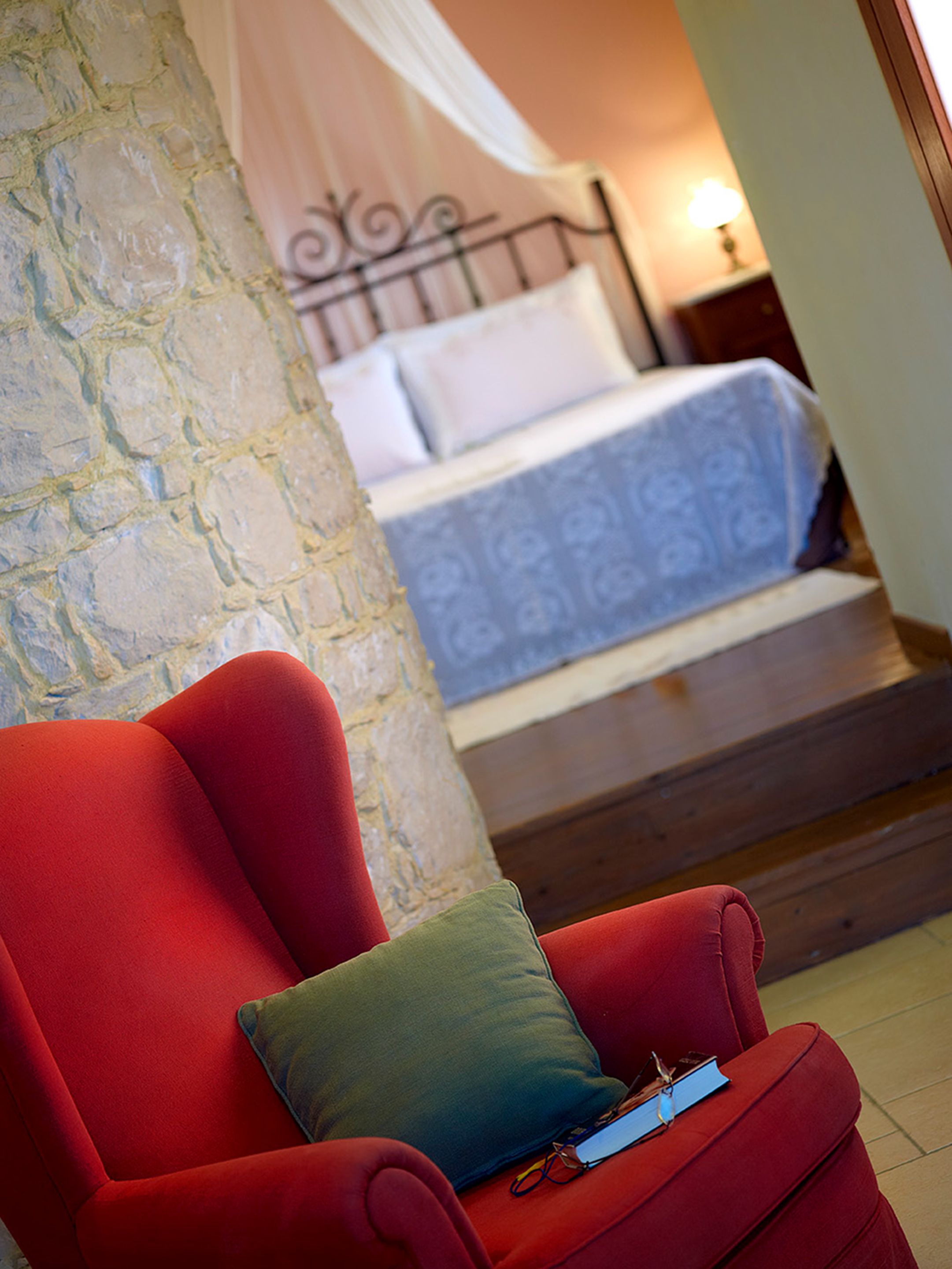 Un fauteuil rouge avec un coussin vert et un livre, un mur en pierre, un lit à baldaquin blanc et une table de chevet avec une lampe.