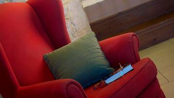 Un fauteuil rouge avec un coussin vert et un livre, un mur en pierre, un lit à baldaquin blanc et une table de chevet avec une lampe.