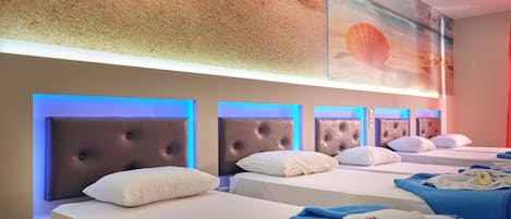 Habitación de hotel con cuatro camas, cada una con cabecero capitonado y ropa de cama blanca. La habitación tiene un mural con tema de playa en la pared y una tira de luz ambiental azul sobre las camas.