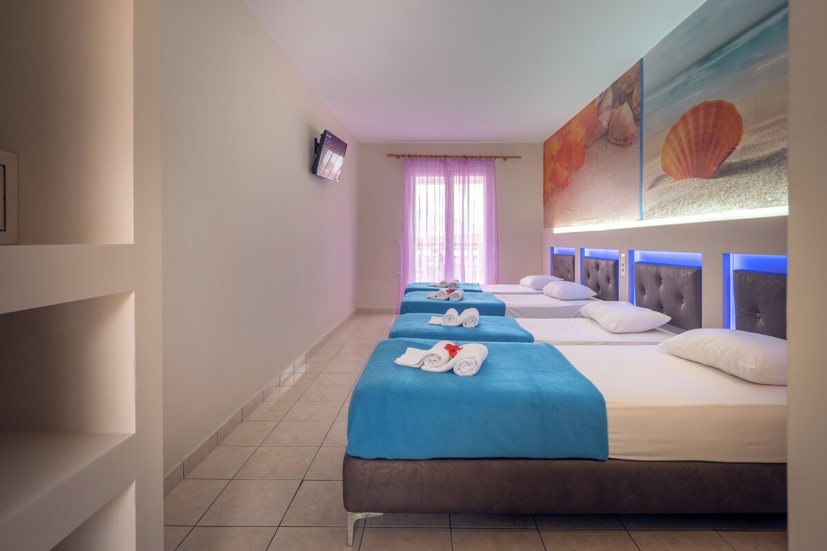 A hotel room with three beds, a TV, a painting of seashells, and a view of the sea through the window.