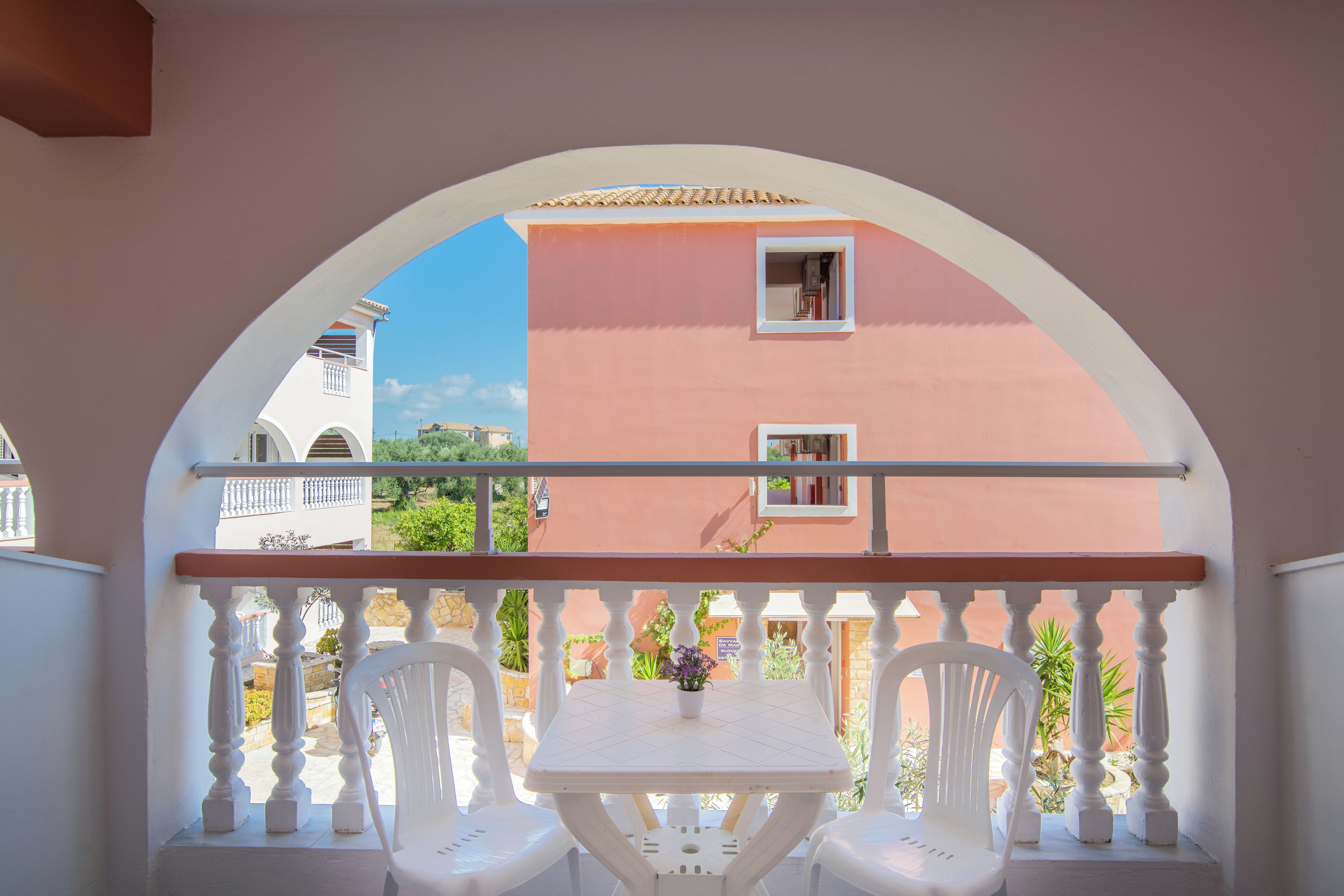 A balcony with a white table and two chairs, a pink building in the background, and a clear blue sky.
