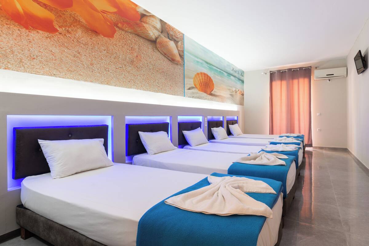 A hotel room with four beds arranged in a row, each with a headboard and a blue blanket.