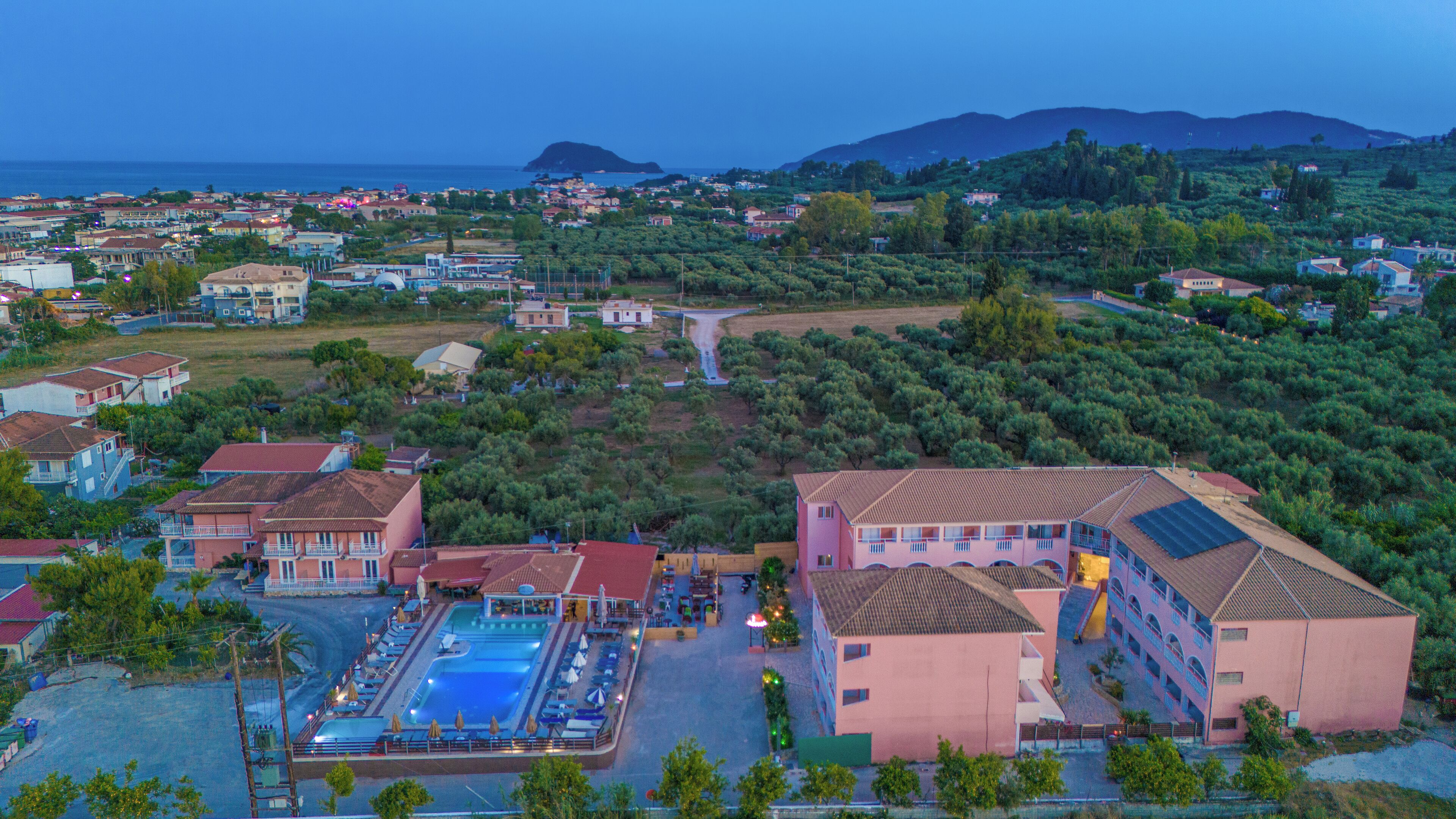 An aerial view of a coastal resort with a swimming pool, multiple buildings, and a view of the sea and mountains in the background.