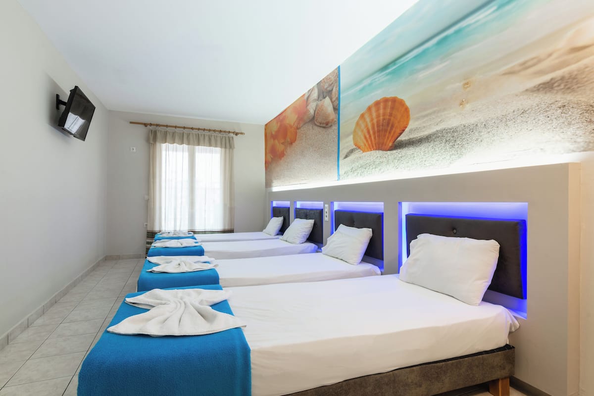 A hotel room with four beds, each with a blue bedspread and white pillows. There is a large painting of seashells above the beds.