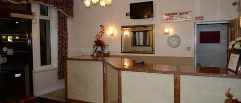 A hotel reception area with a marble counter, a chandelier, a television, a warning notice, a clock, and a mirror.