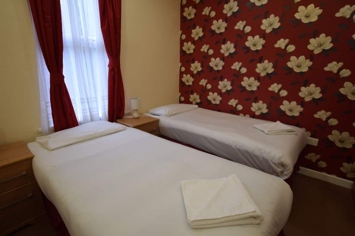 Habitación de hotel con dos camas individuales, una mesita de noche de madera, un papel tapiz floral rojo y una ventana con cortinas rojas.