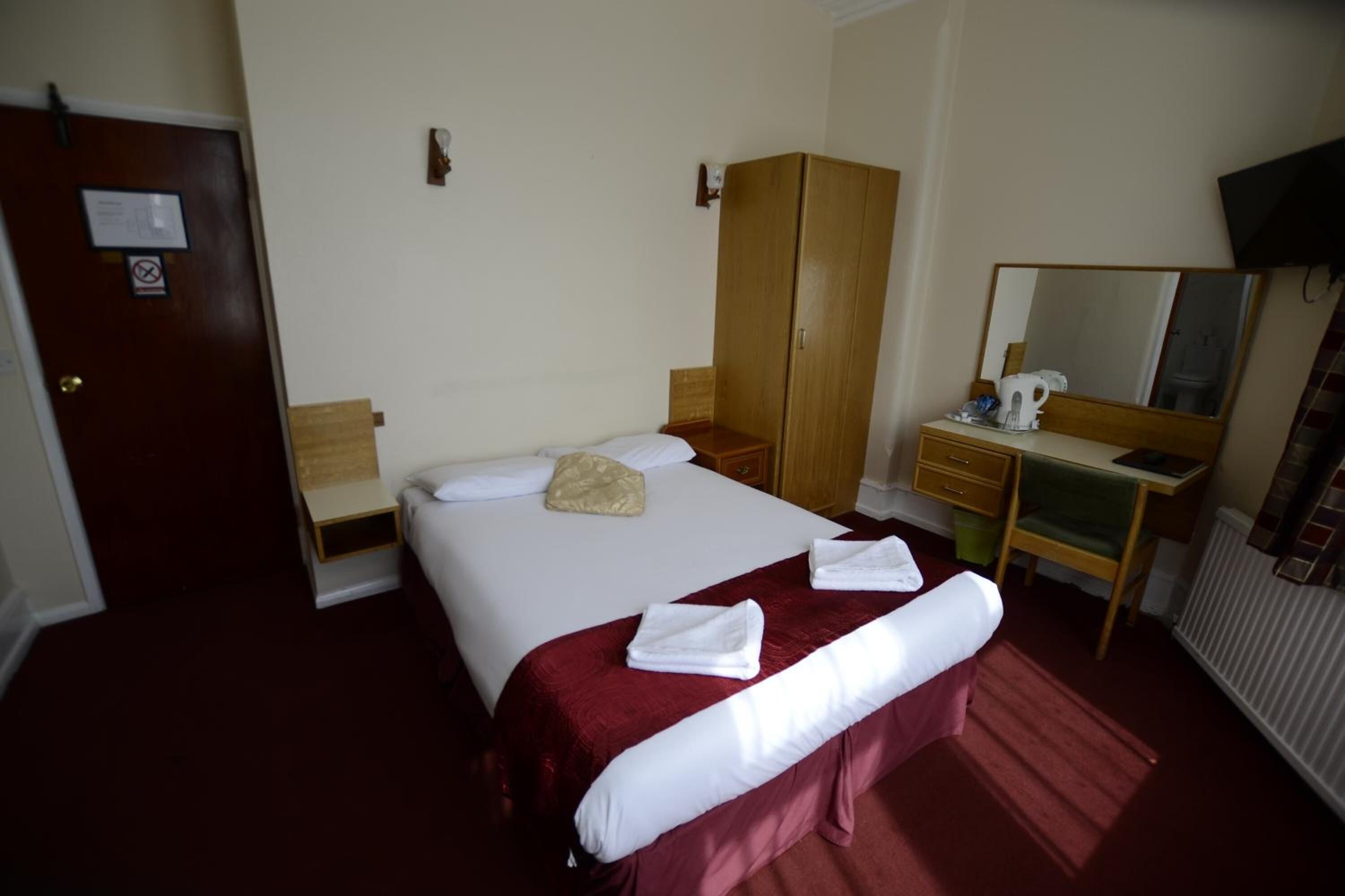 Une chambre d’hôtel comprenant un lit, une armoire en bois, un bureau avec une chaise, un miroir et un radiateur.