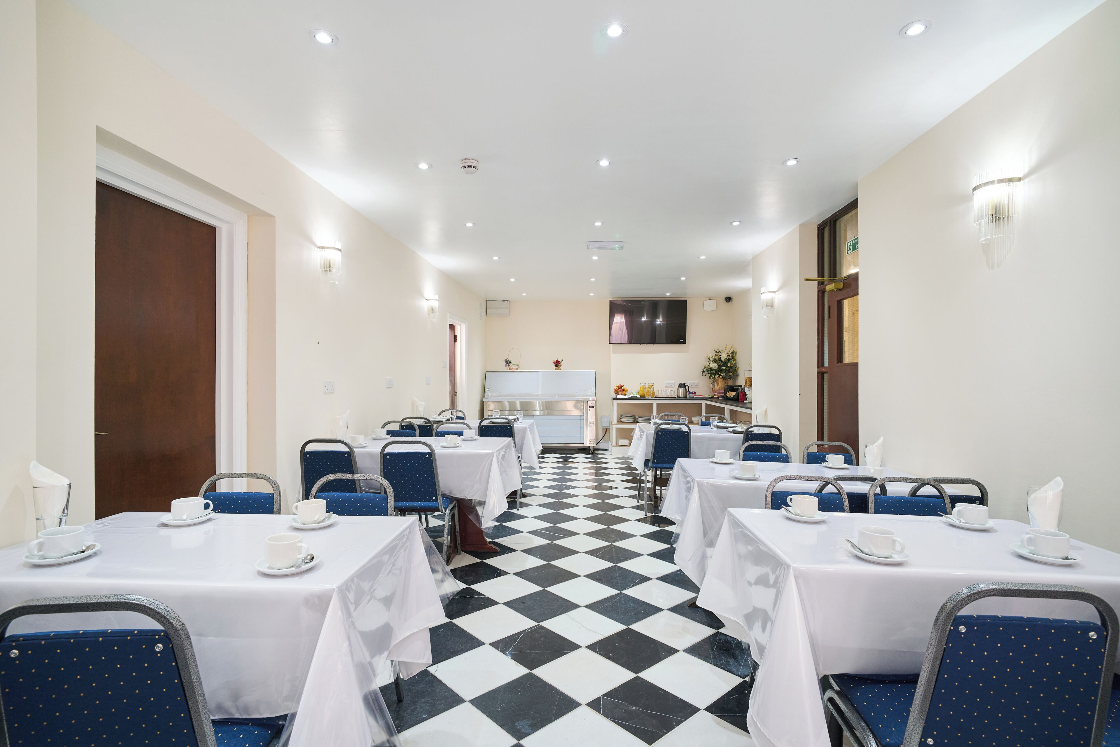 Un comedor de hotel con mesas y sillas, suelo a cuadros y una cocina al fondo.