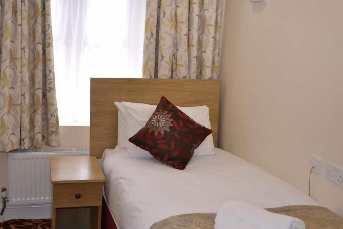 A neatly made bed with a pillow, a bedside table, a radiator, and curtains.