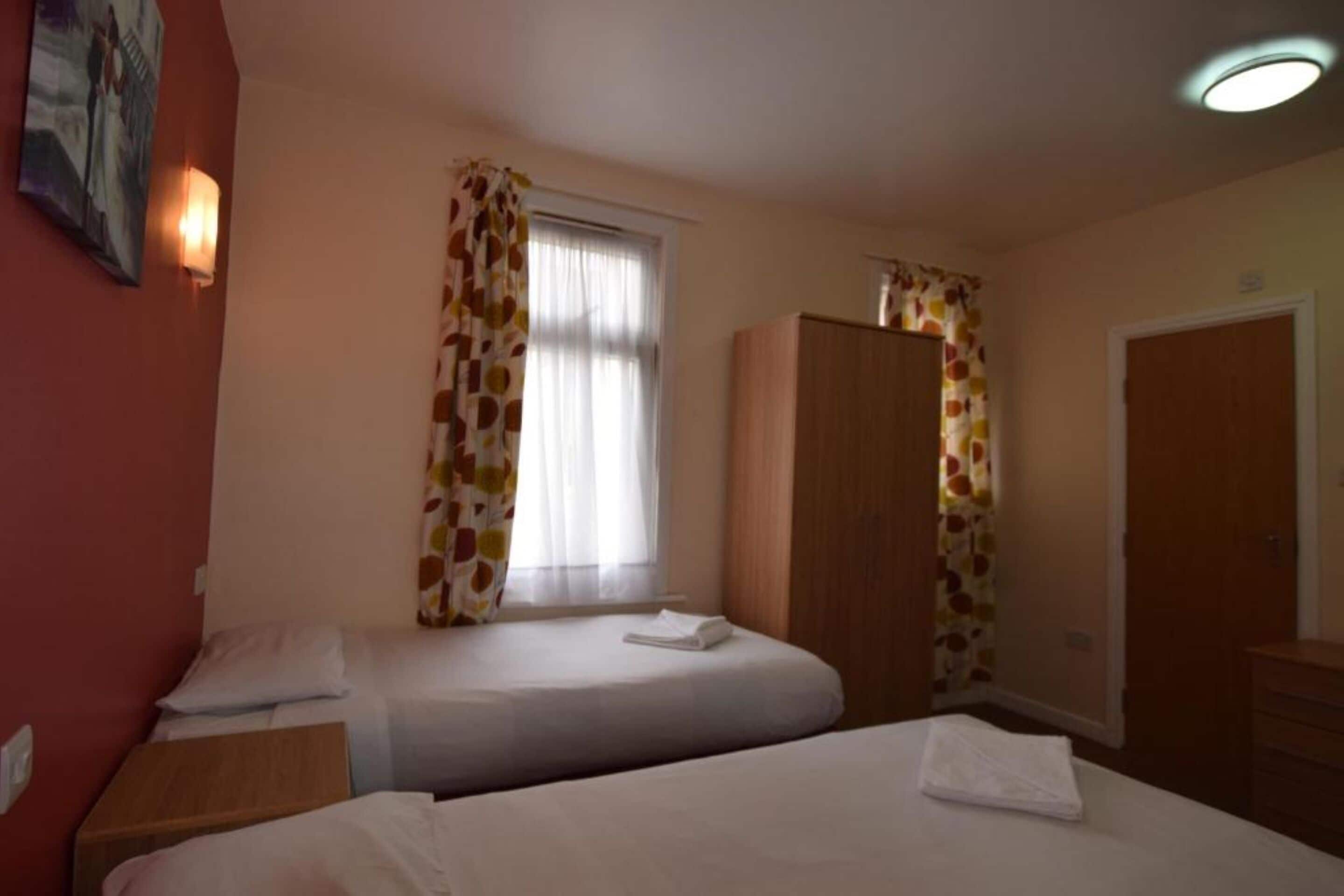A hotel room with two beds, a wardrobe, a window with curtains, and a door.