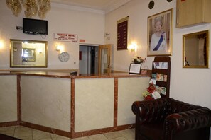 Zona de recepción de un hotel con mostrador de mármol, sofá de cuero, televisor, un aviso de precaución, un reloj y un cuadro en la pared.