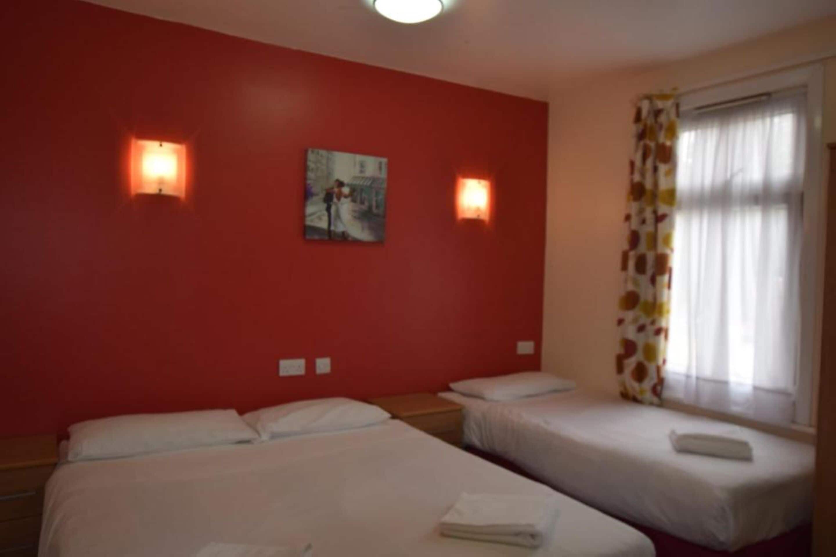 A hotel room with two beds, a red wall, and two wall-mounted lights.