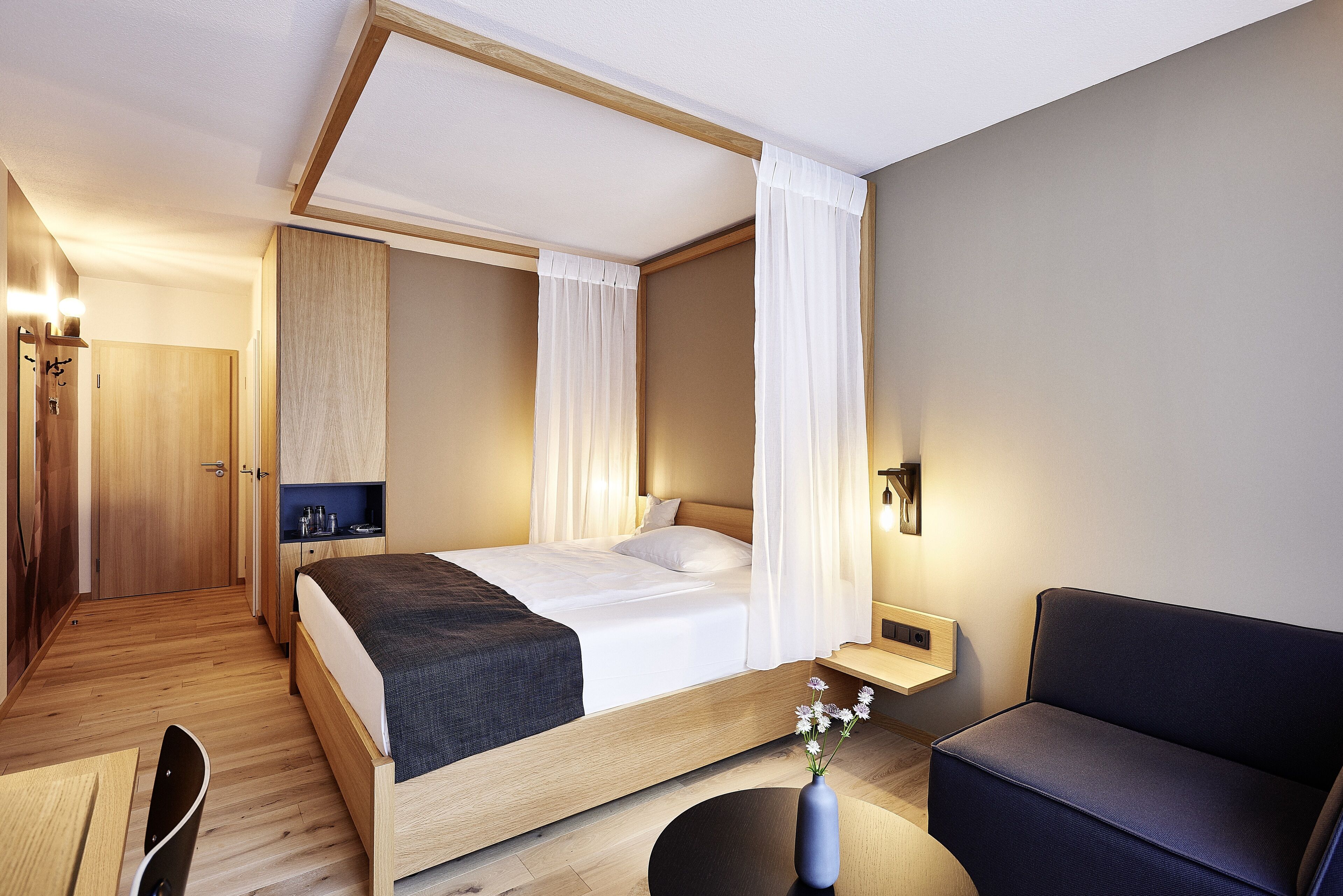 Quarto de hotel moderno com uma cama grande, uma escrivaninha, uma cadeira e uma pequena mesa com um vaso de flores.