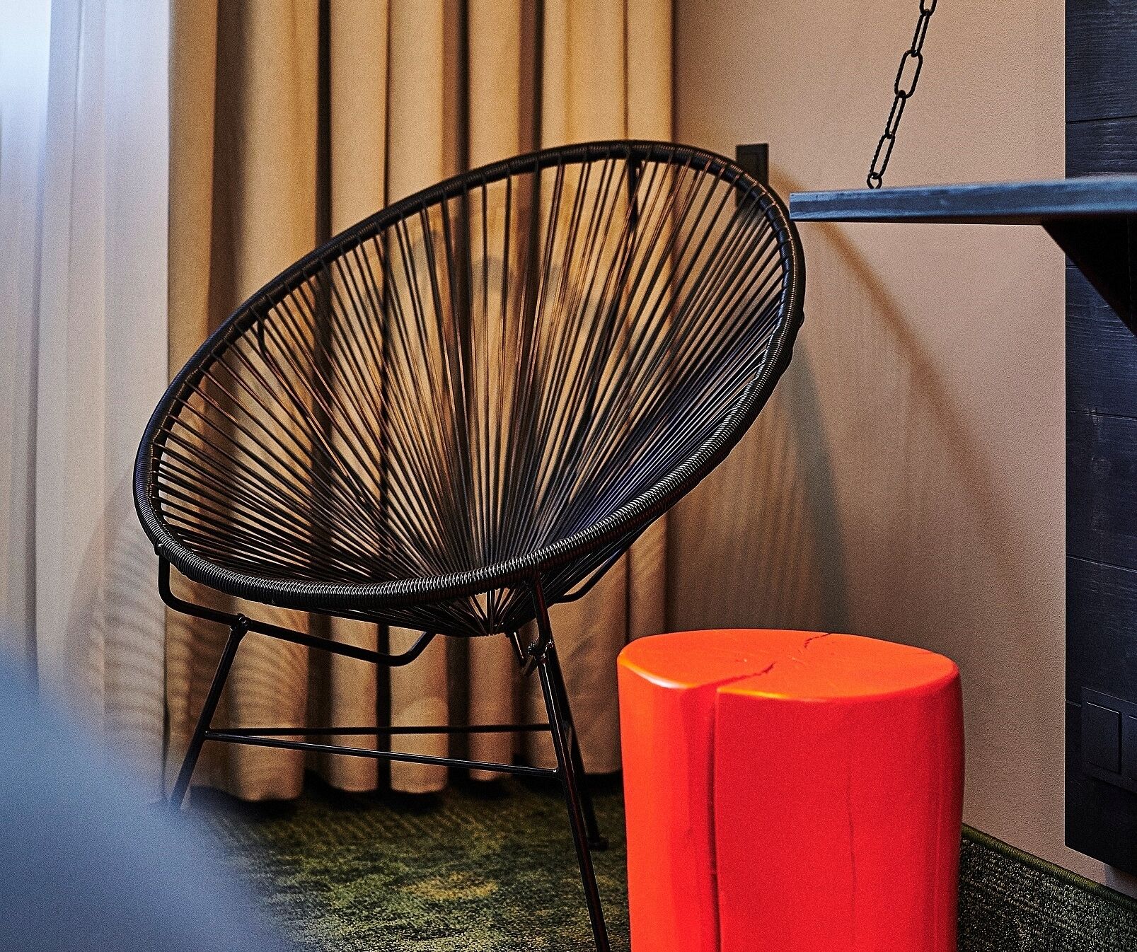 A black wireframe chair and a red stool in a room with beige curtains and a black wall.