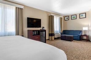A hotel room with a large bed, a flat-screen TV, a minibar, a sofa, and a small table with a lamp. - Comfort Suites Plano - Dallas North (Plano)