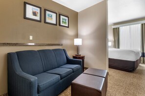 A hotel room with a dark blue sofa, a small table, a lamp, and a bed with a white comforter. There are three framed pictures on the wall above the sofa. - Comfort Suites Plano - Dallas North (Plano)