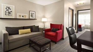 A hotel room with a sofa, armchair, table, and a small ottoman. There are framed pictures on the wall and a lamp on a side table. - Country Inn & Suites by Radisson, Kansas City at Village West, KS (Kansas City)