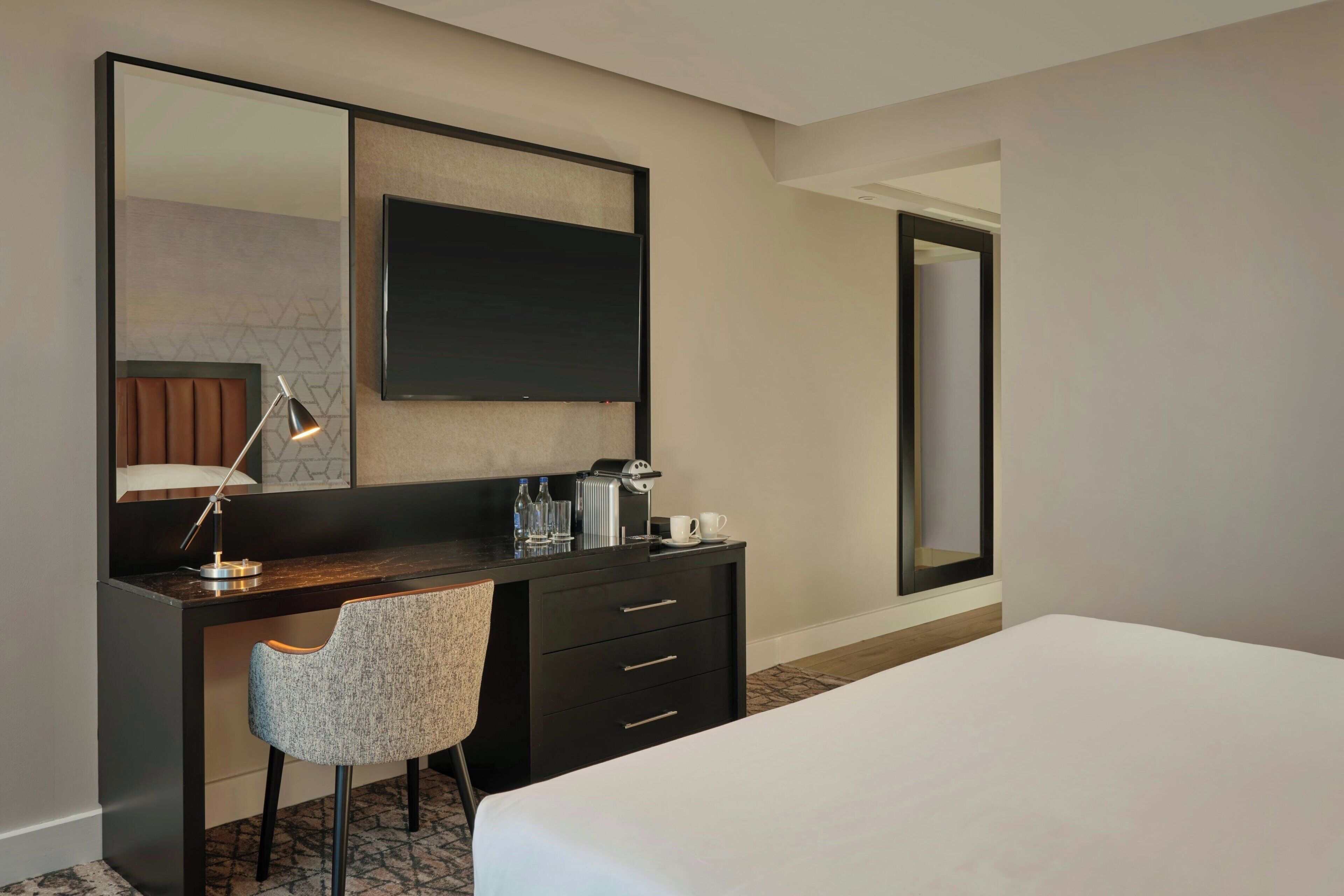 Habitación de hotel con cama, escritorio con silla, espejo grande, televisor de pantalla plana y cafetera.