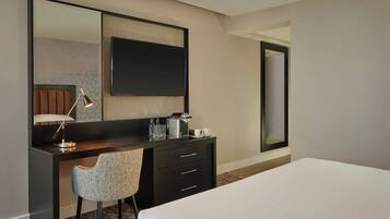 Habitación de hotel con cama, escritorio con silla, espejo grande, televisor de pantalla plana y cafetera.