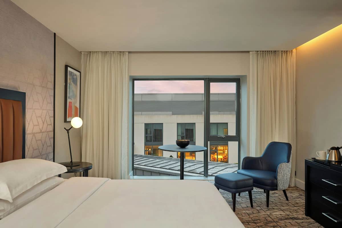 A hotel room with a bed, a small table with a bowl, a chair, a dresser, and a view of the building outside.
