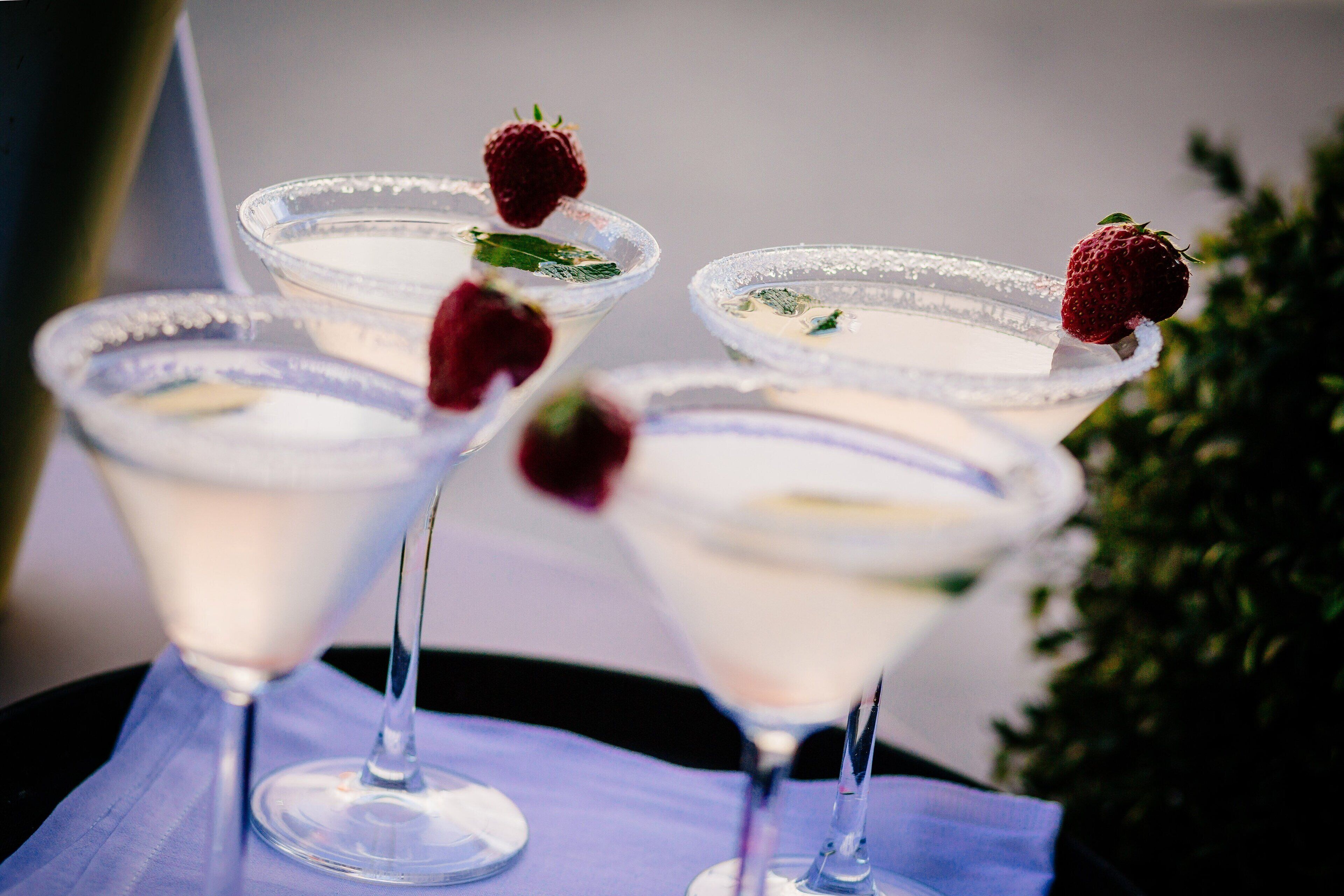 Four martini glasses with a clear liquid, garnished with strawberries, on a table with a blue cloth.