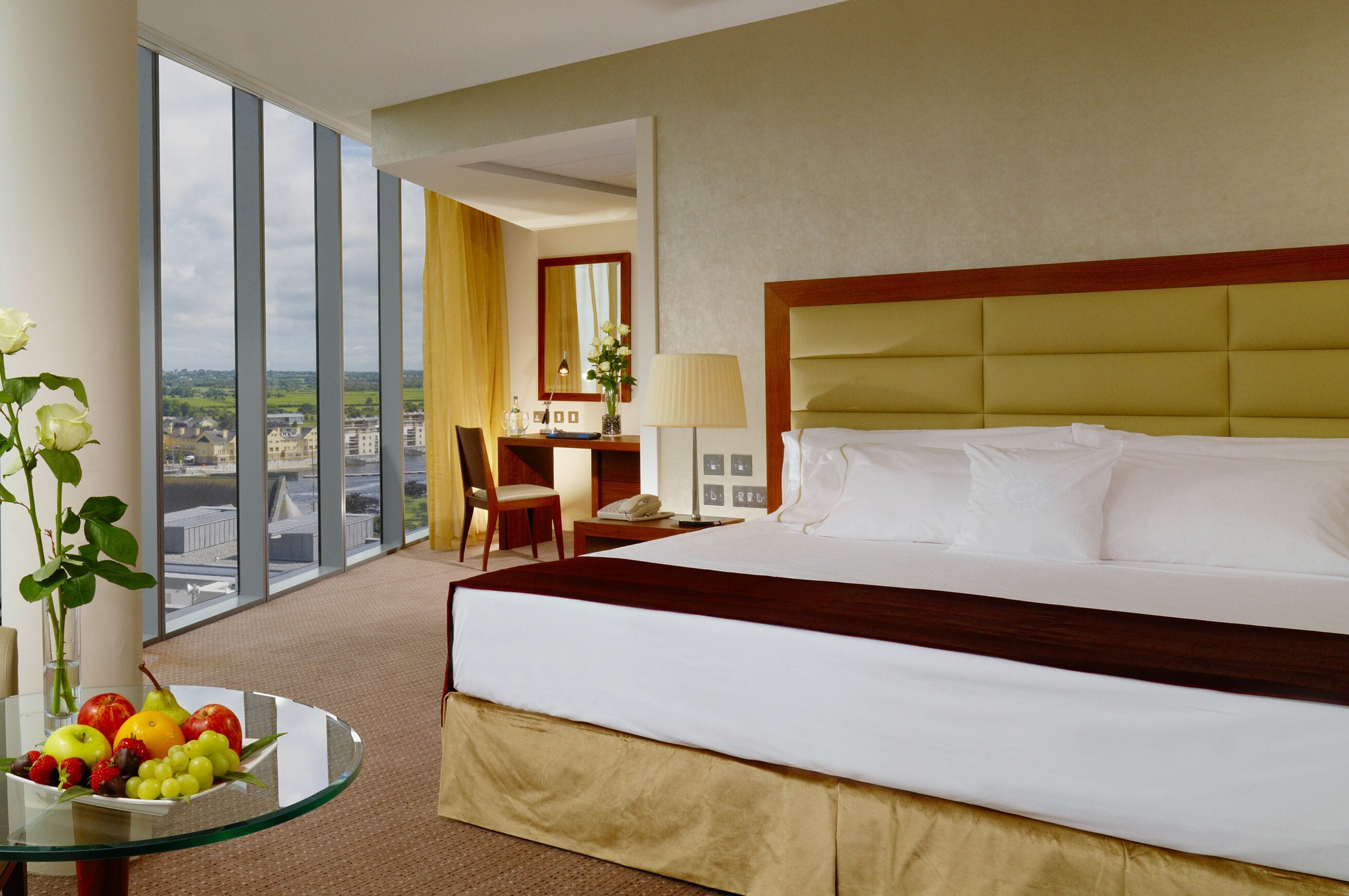 A hotel room with a large bed, a desk with a lamp, a chair, and a view of the city through large windows.