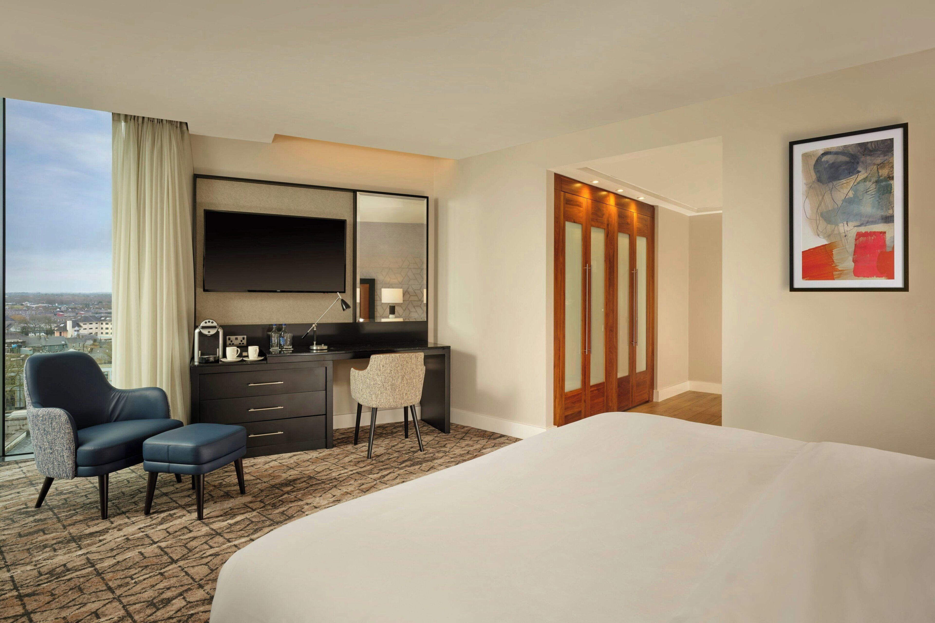 A hotel room with a large bed, a desk with a chair, a TV, and a city view.
