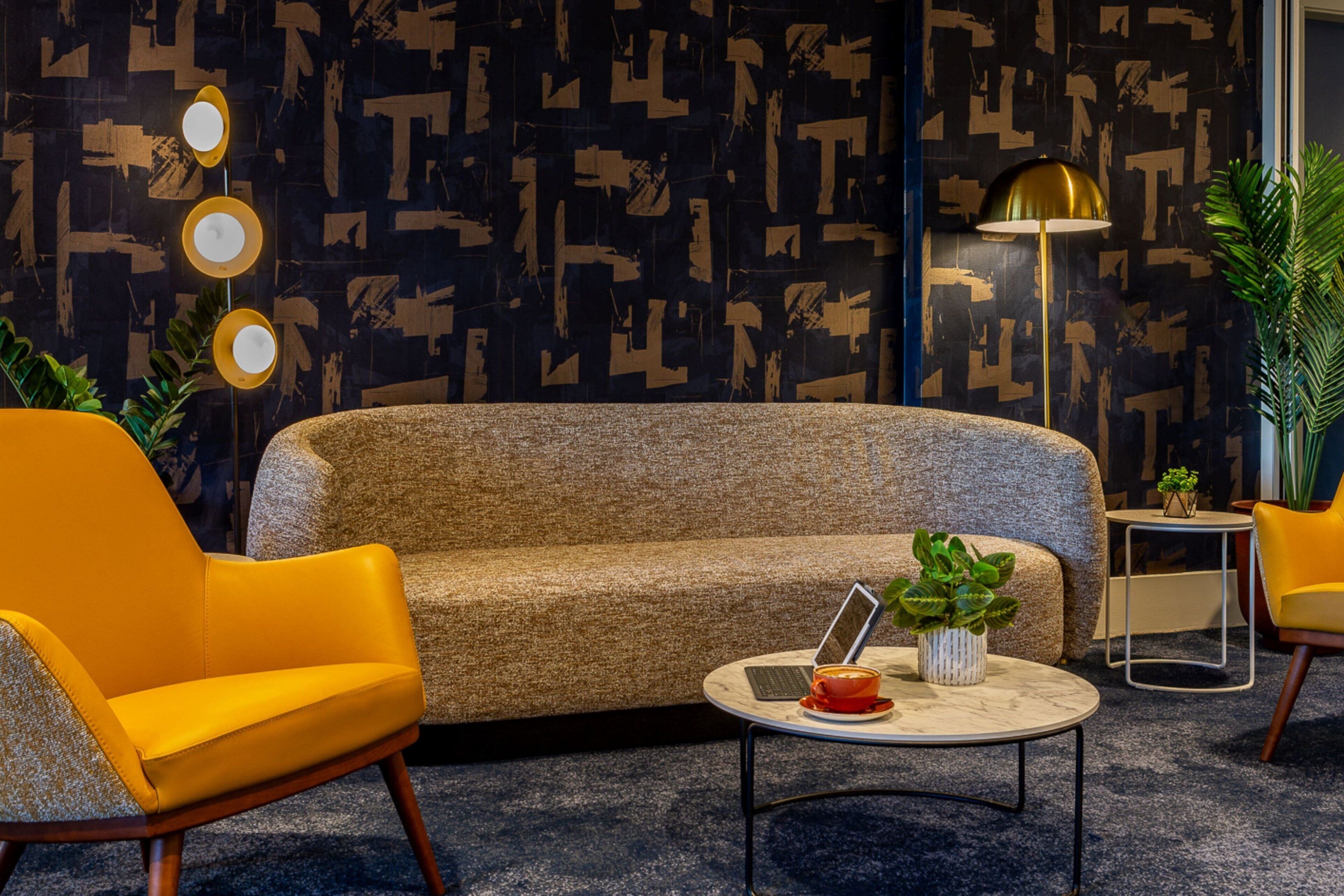 Een moderne woonkamer met een bank, twee gele fauteuils, een salontafel met een laptop en een kopje, en een vloerlamp.