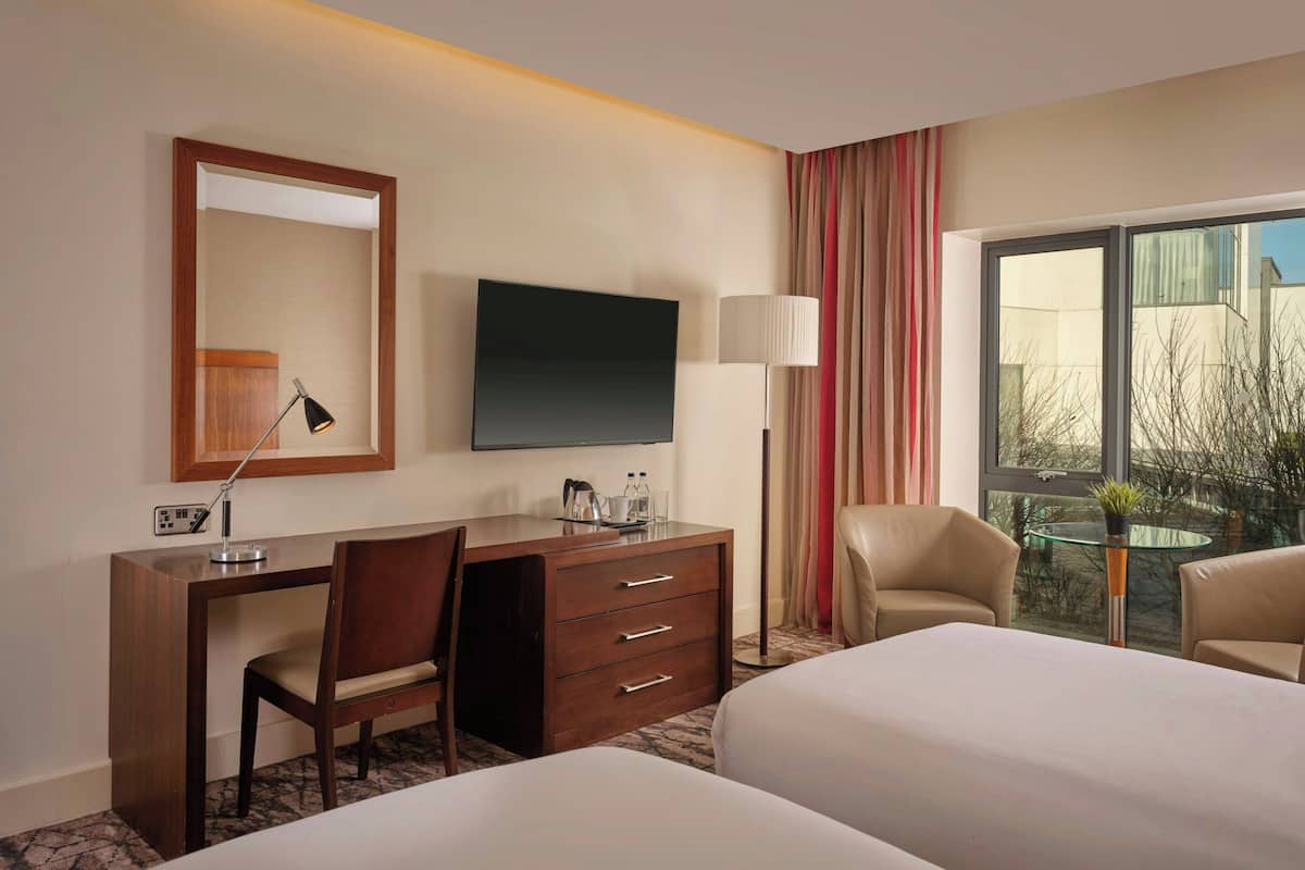 A hotel room with a desk, chair, TV, two beds, a lamp, and a view of the outdoors.