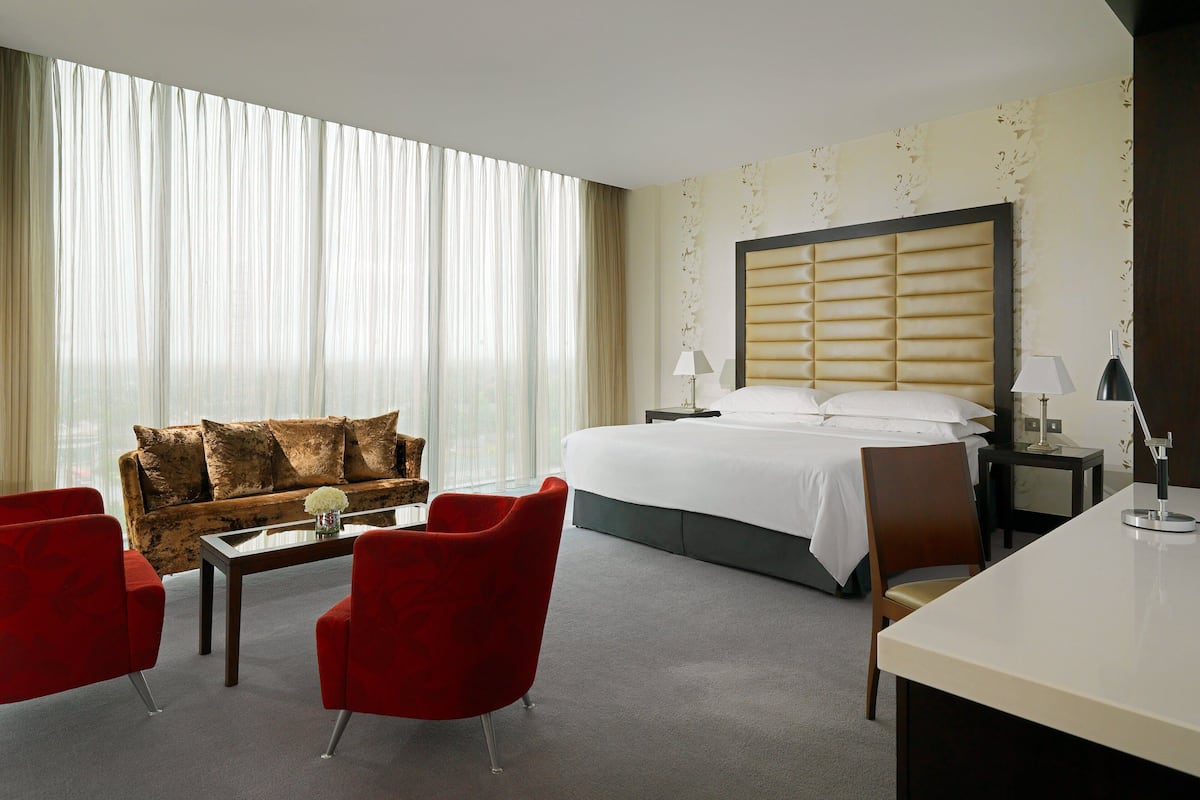 A hotel room with a large bed, a sofa, two red chairs, a desk, and a nightstand with a lamp.