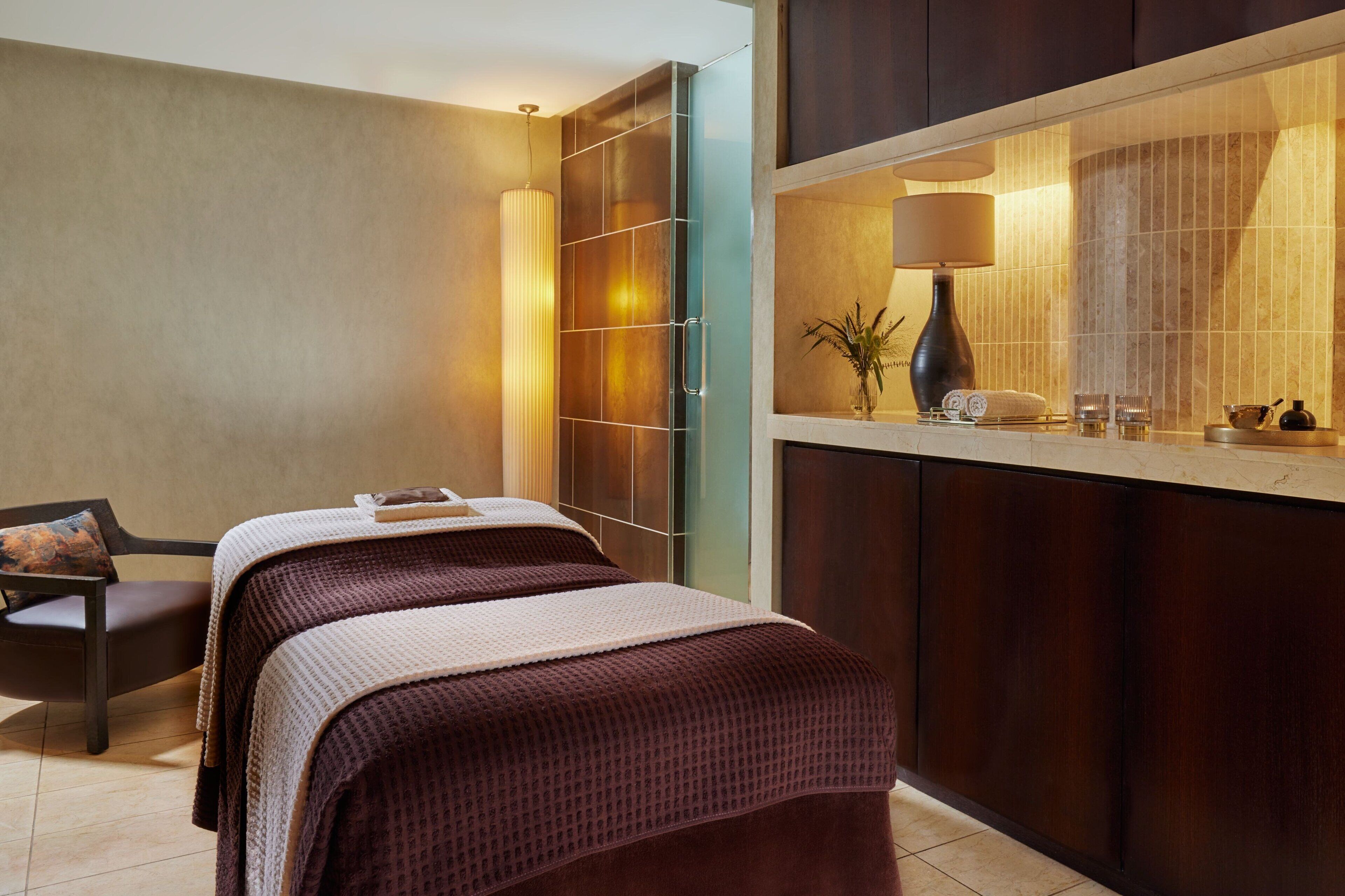 A hotel room with two beds, a sitting area with a chair and a table, a large mirror, and a minibar.