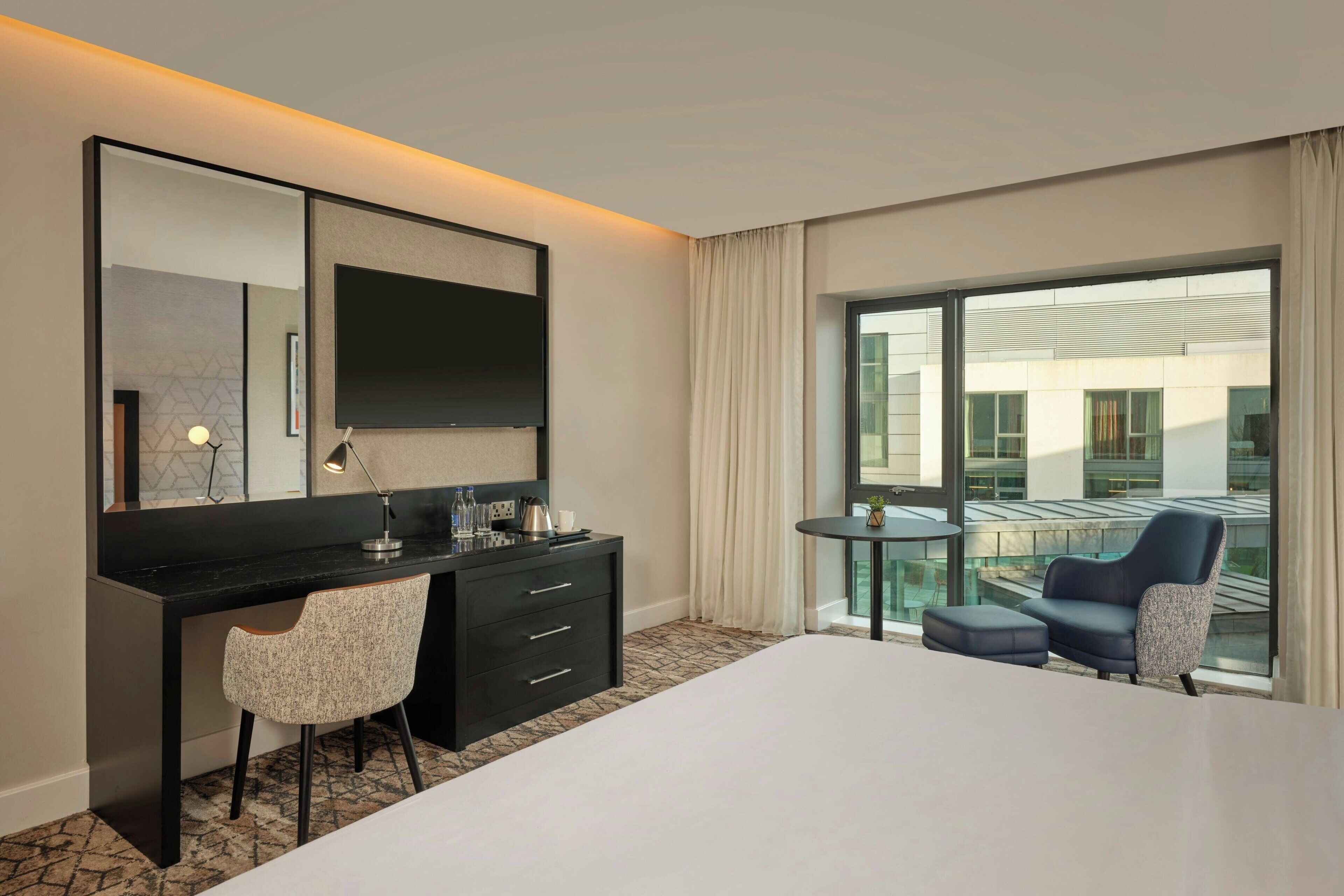 A hotel room with a large bed, a desk with a chair, a small table, a sofa, and a large window offering a view of the building's exterior.