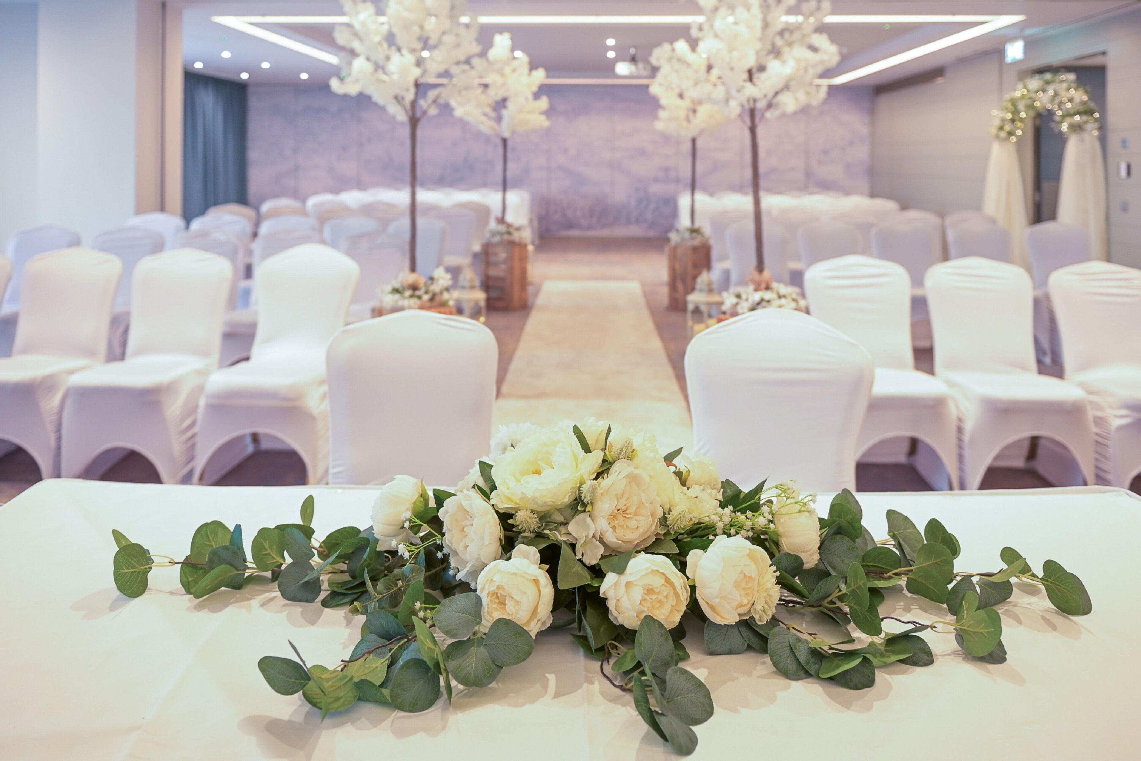 Eine Hochzeitslocation mit Reihen weißer Stühle, einer floralen Tischdekoration und einer neutralen Wand im Hintergrund.